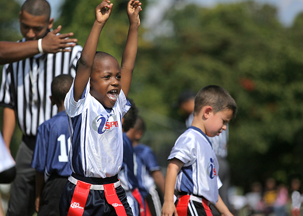 pee wee age group male flag football player raises both arms in celebration of scoring a touchdown as a referee looks on and a teammate walks away.