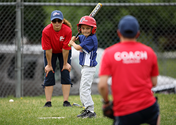 i9 Sports baseball player is standing in the batters box awaiting a pitch to be thrown by the coach as another coach watches from behind home plate